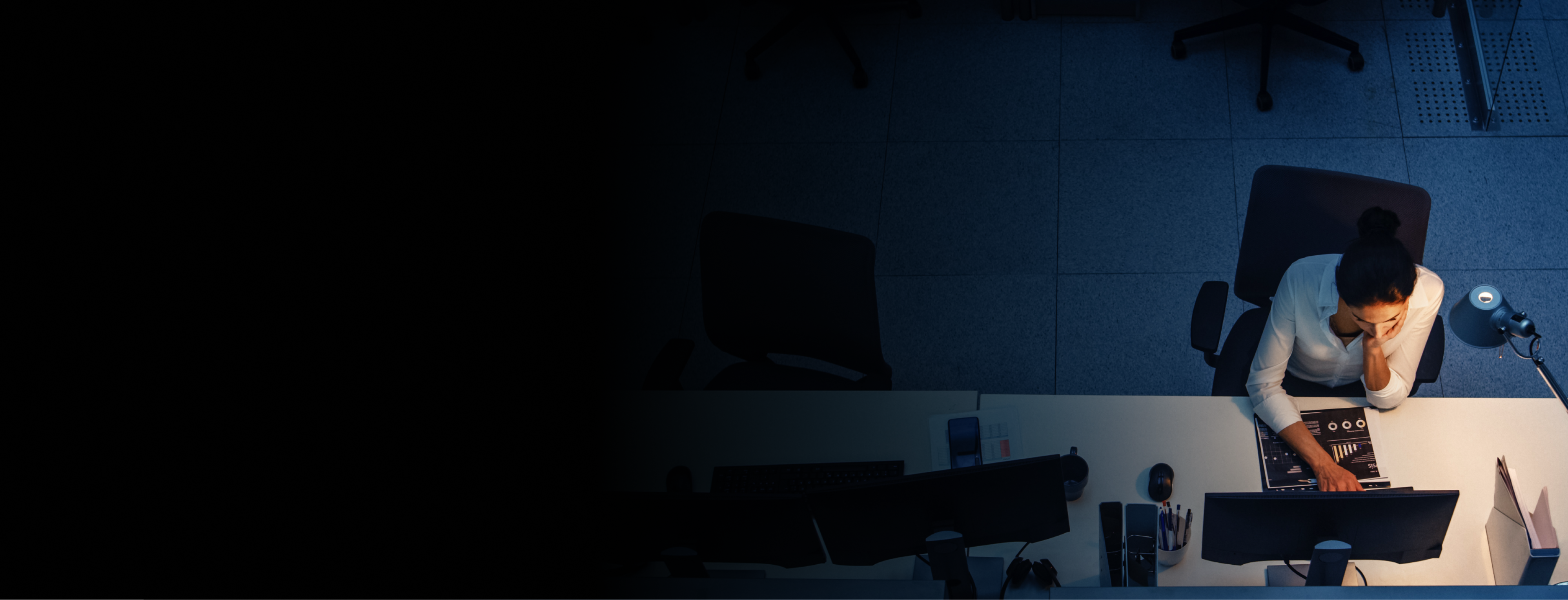 Woman on a computer in a dark room. Lit desk, dark blue background. 